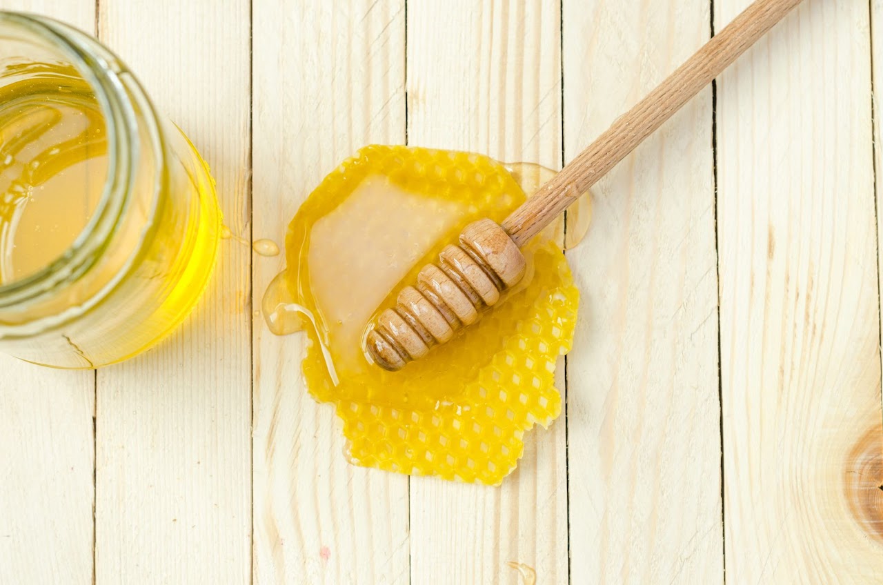 A Wooden Honey Spoon Laying on a Puddle of Honey Next to a Honey Jar